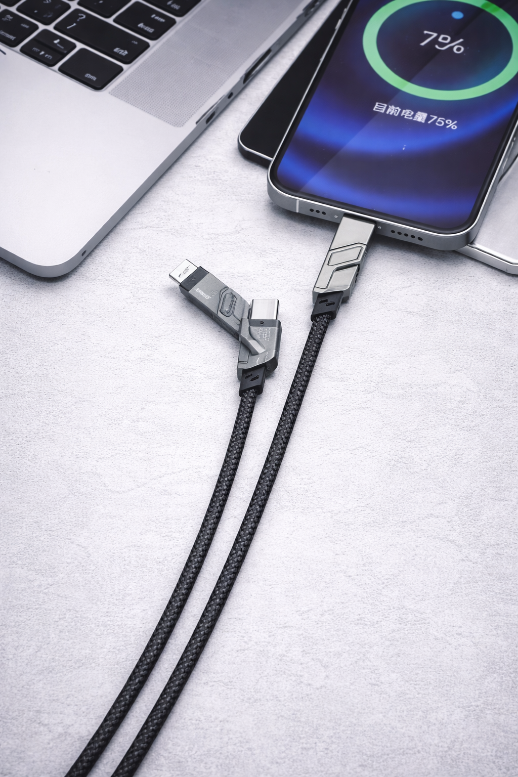 Black braided cable charging a smartphone with a laptop in the background on a light gray surface.